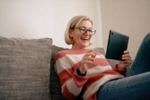 mature blonde woman sitting on sofa and using tablet for video call - WorldTone